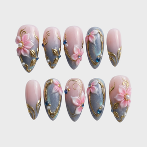 Pink and grey gradient press-on nails featuring 3D floral art, gold metallic swirls, pearls, and blue rhinestones.