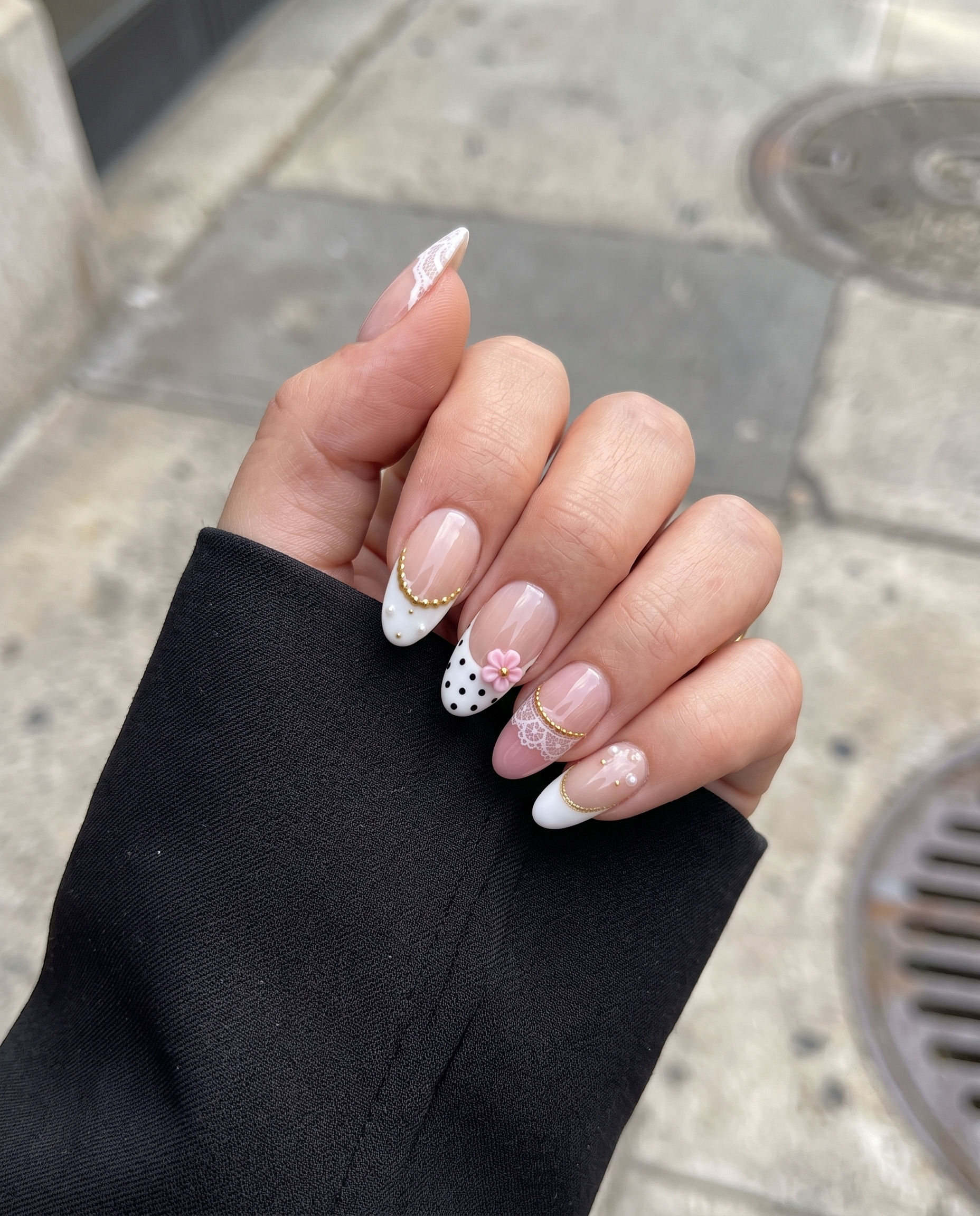 A set of press-on nails with white lace patterns, black polka dots, 3D pink flowers, gold beads, and pearl embellishments.