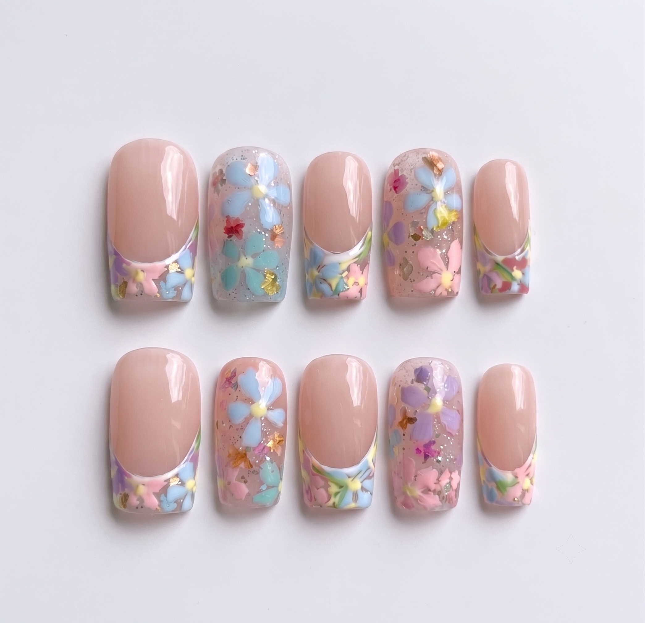 A set of press-on nails featuring a French tip design with floral motifs, gold foil, and glitter on a sheer base.