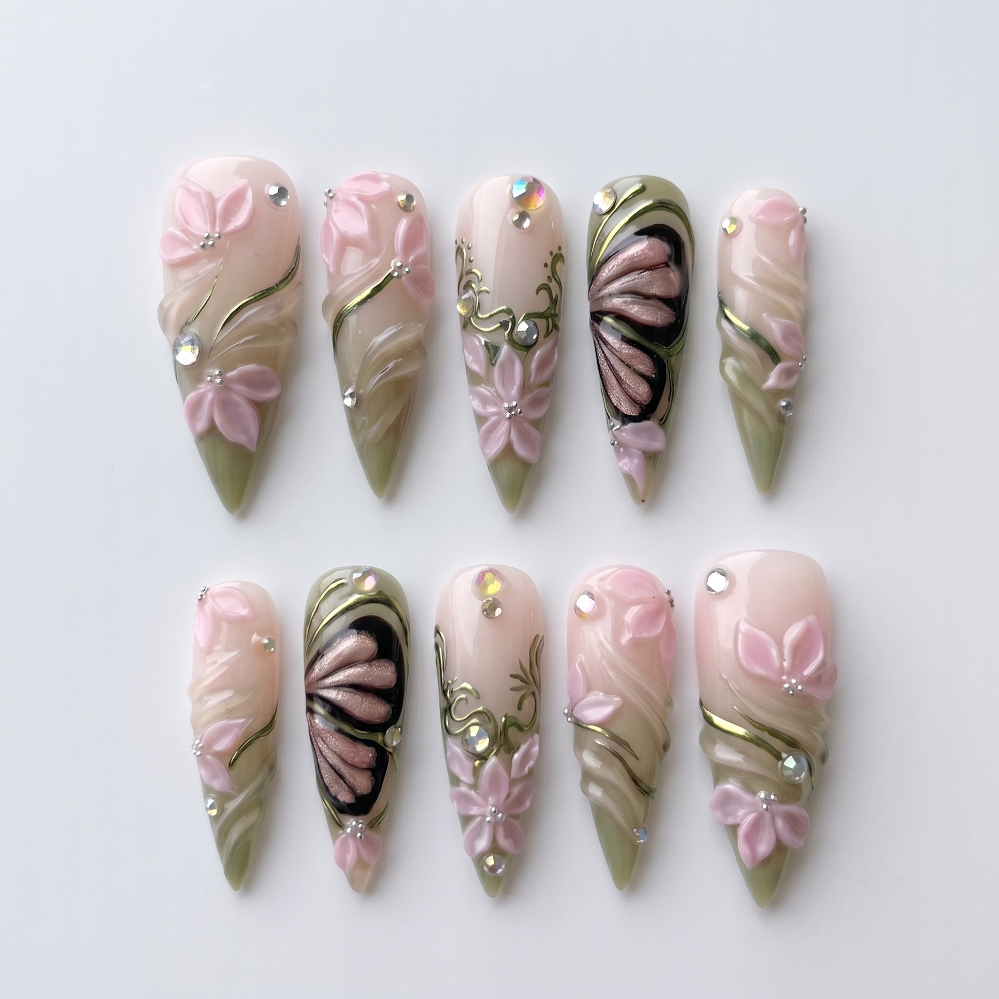 Stiletto press-on nails featuring 3D pink flowers, butterfly wings, sage green swirls, and rhinestones.