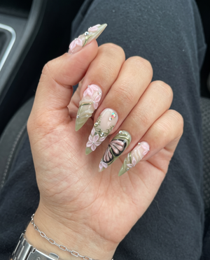 Stiletto press-on nails featuring 3D pink flowers, butterfly wings, sage green swirls, and rhinestones.