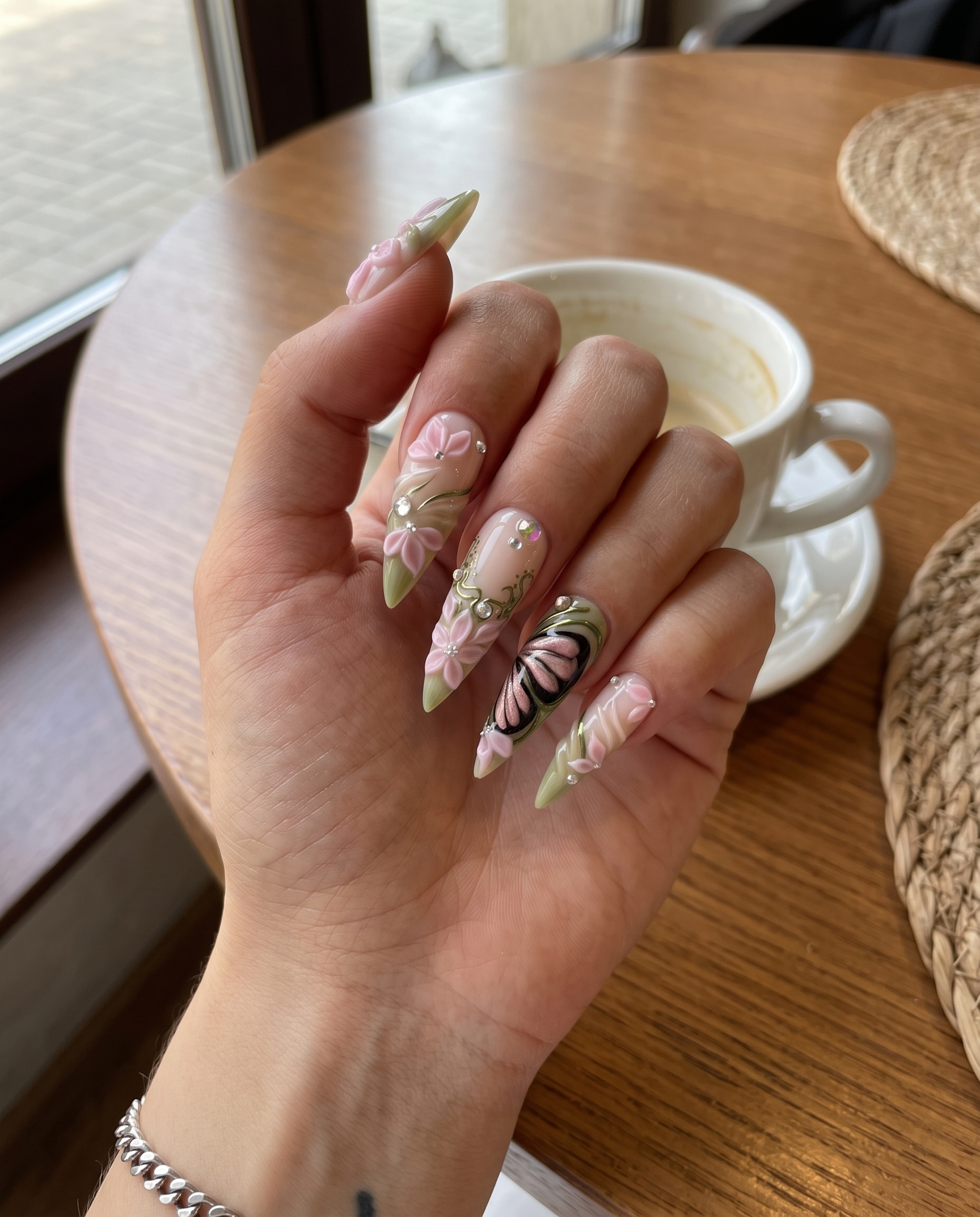 Stiletto press-on nails featuring 3D pink flowers, butterfly wings, sage green swirls, and rhinestones.