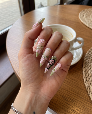 Stiletto press-on nails featuring 3D pink flowers, butterfly wings, sage green swirls, and rhinestones.