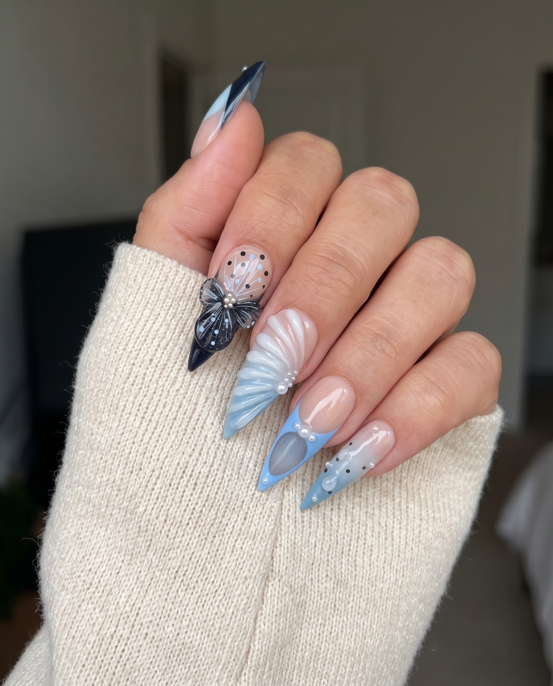 Set of stiletto press-on nails featuring blue and black French tips, butterfly art, pearl accents, and 3D shell textures.