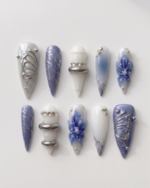 A set of press-on nails featuring blue floral designs, 3D silver chrome butterfly wings, and metallic ring accents.