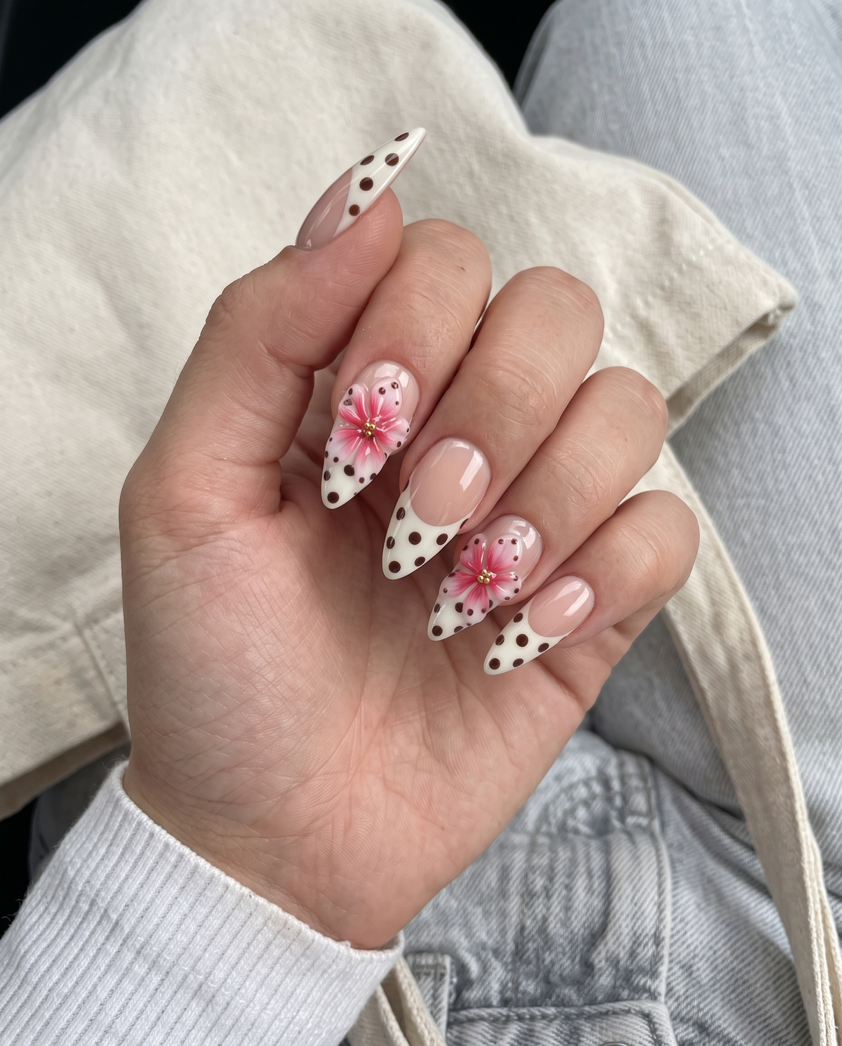 A set of press-on nails featuring a nude base, white French tips with black polka dots, and pink 3D floral accents.