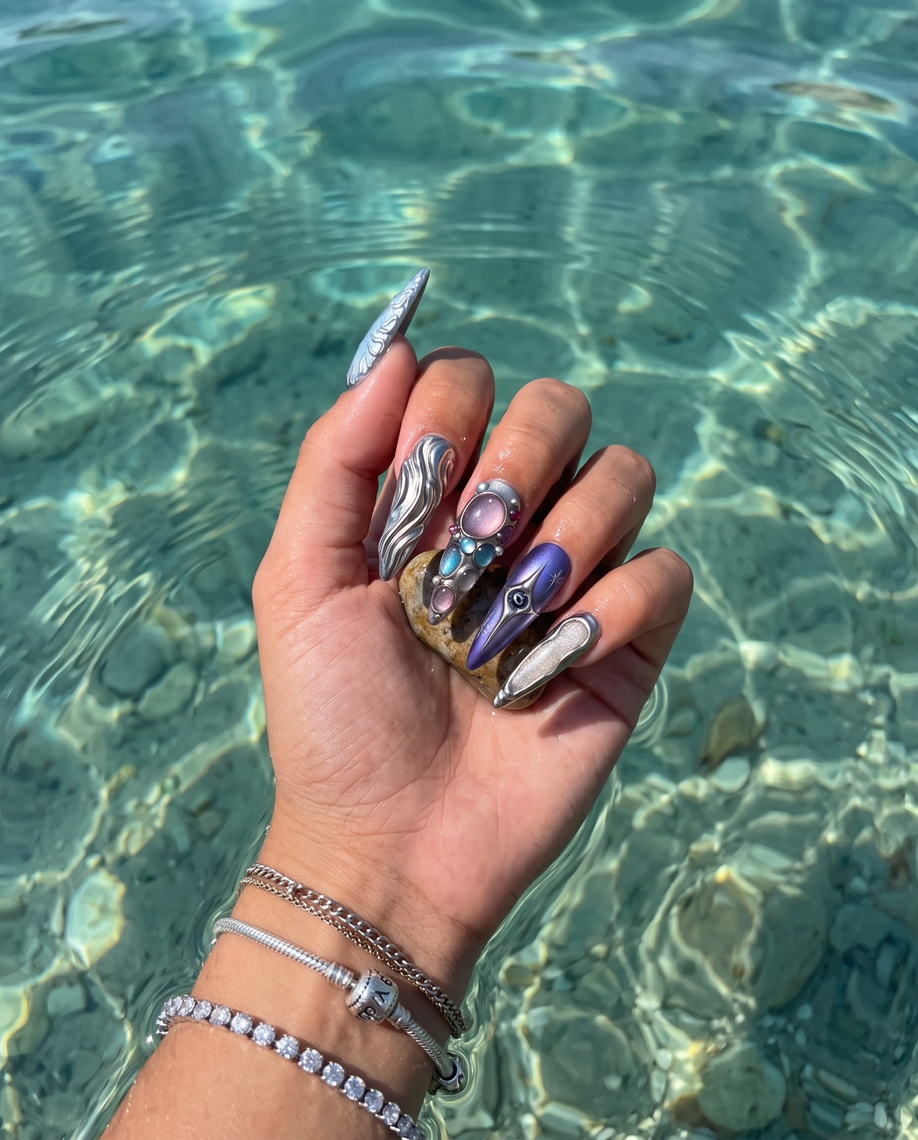 A set of ten stiletto-shaped press-on nails with silver metallic liquid textures, celestial star designs, and embedded colorful gemstones.