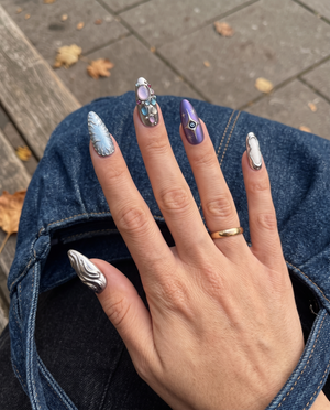 A set of ten stiletto-shaped press-on nails with silver metallic liquid textures, celestial star designs, and embedded colorful gemstones.