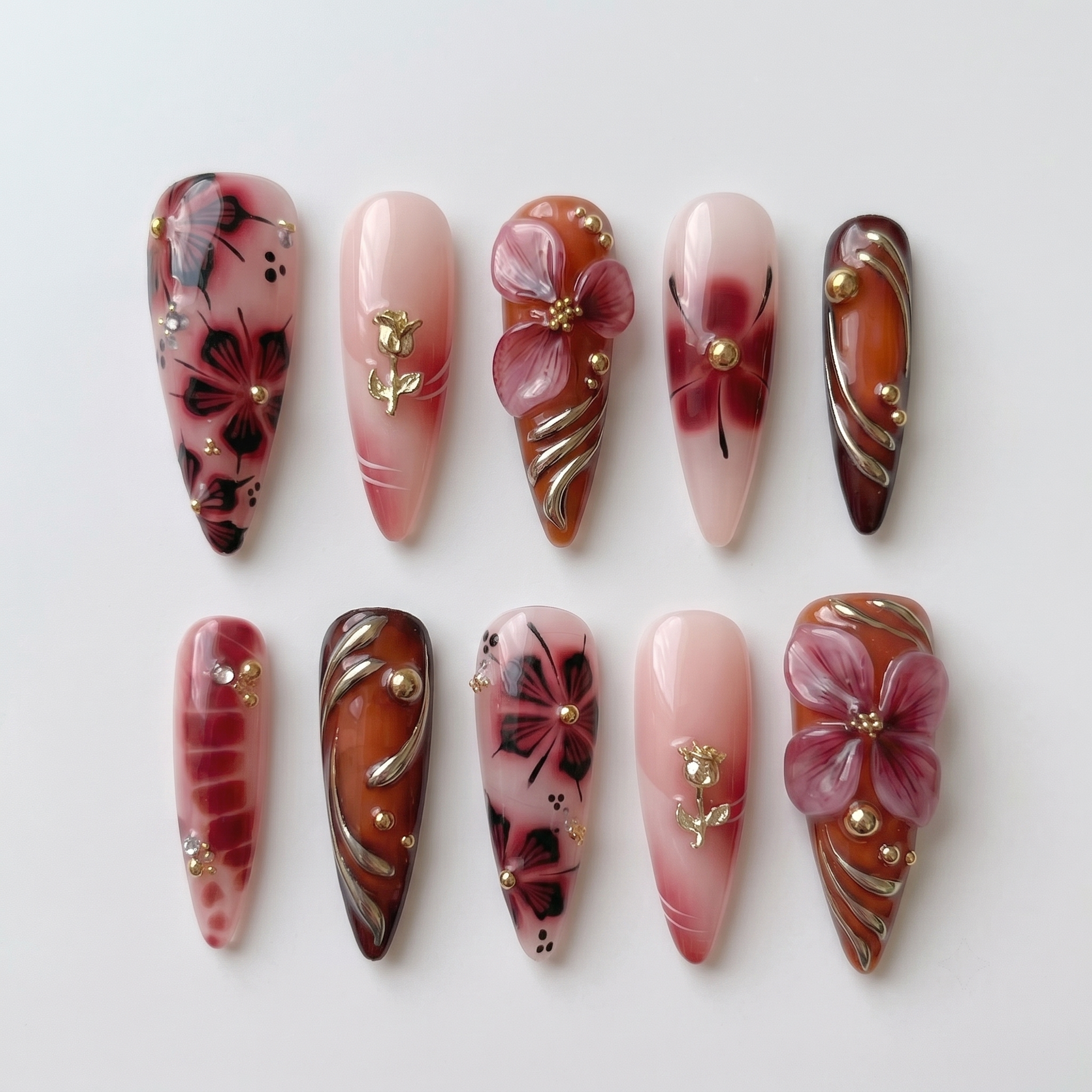 Stiletto shaped press-on nails featuring burgundy and nude polish with 3D floral accents, gold rose charms, and metallic swirl designs.