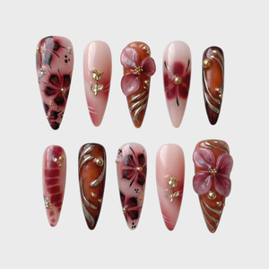 Stiletto shaped press-on nails featuring burgundy and nude polish with 3D floral accents, gold rose charms, and metallic swirl designs.