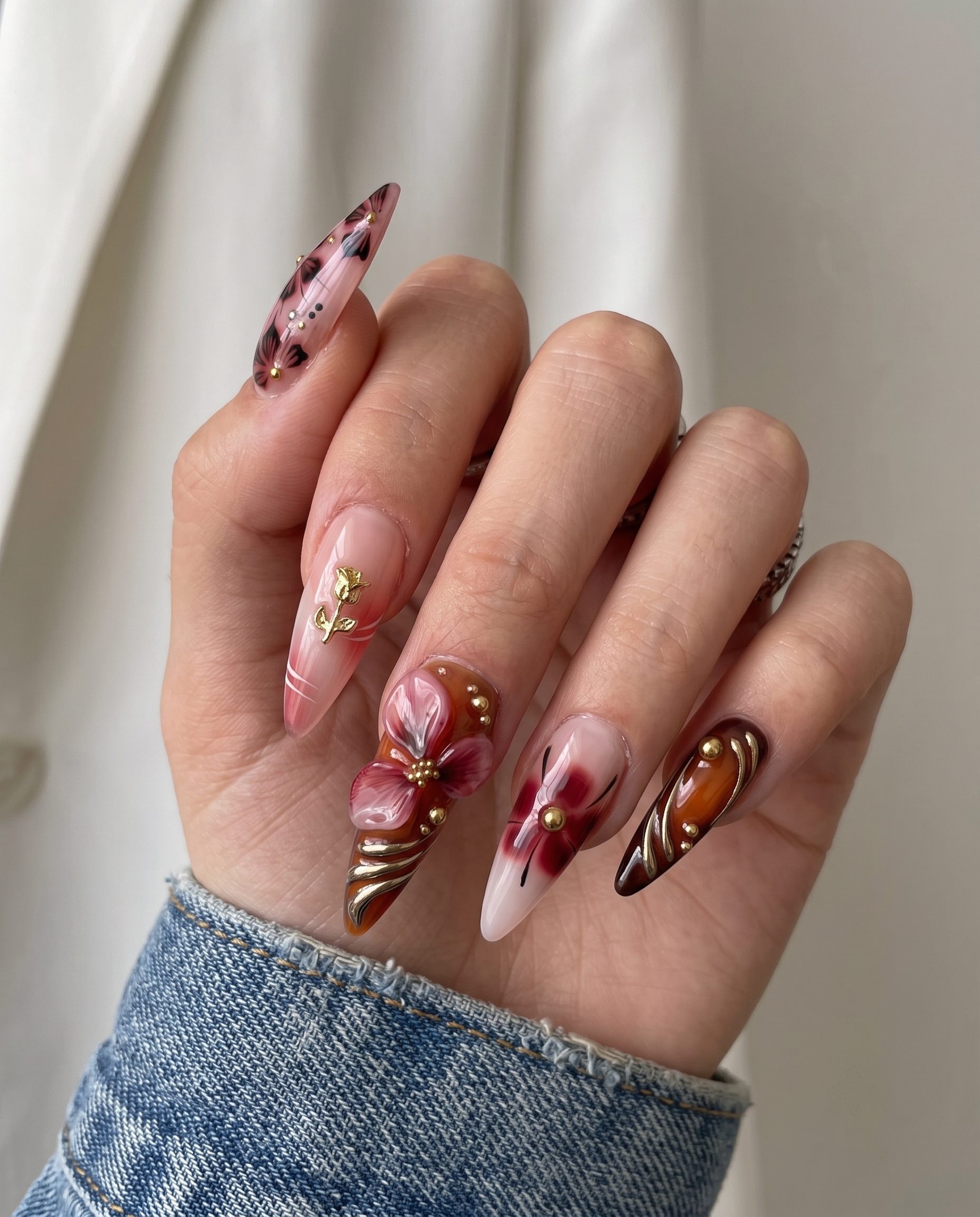 Stiletto shaped press-on nails featuring burgundy and nude polish with 3D floral accents, gold rose charms, and metallic swirl designs.