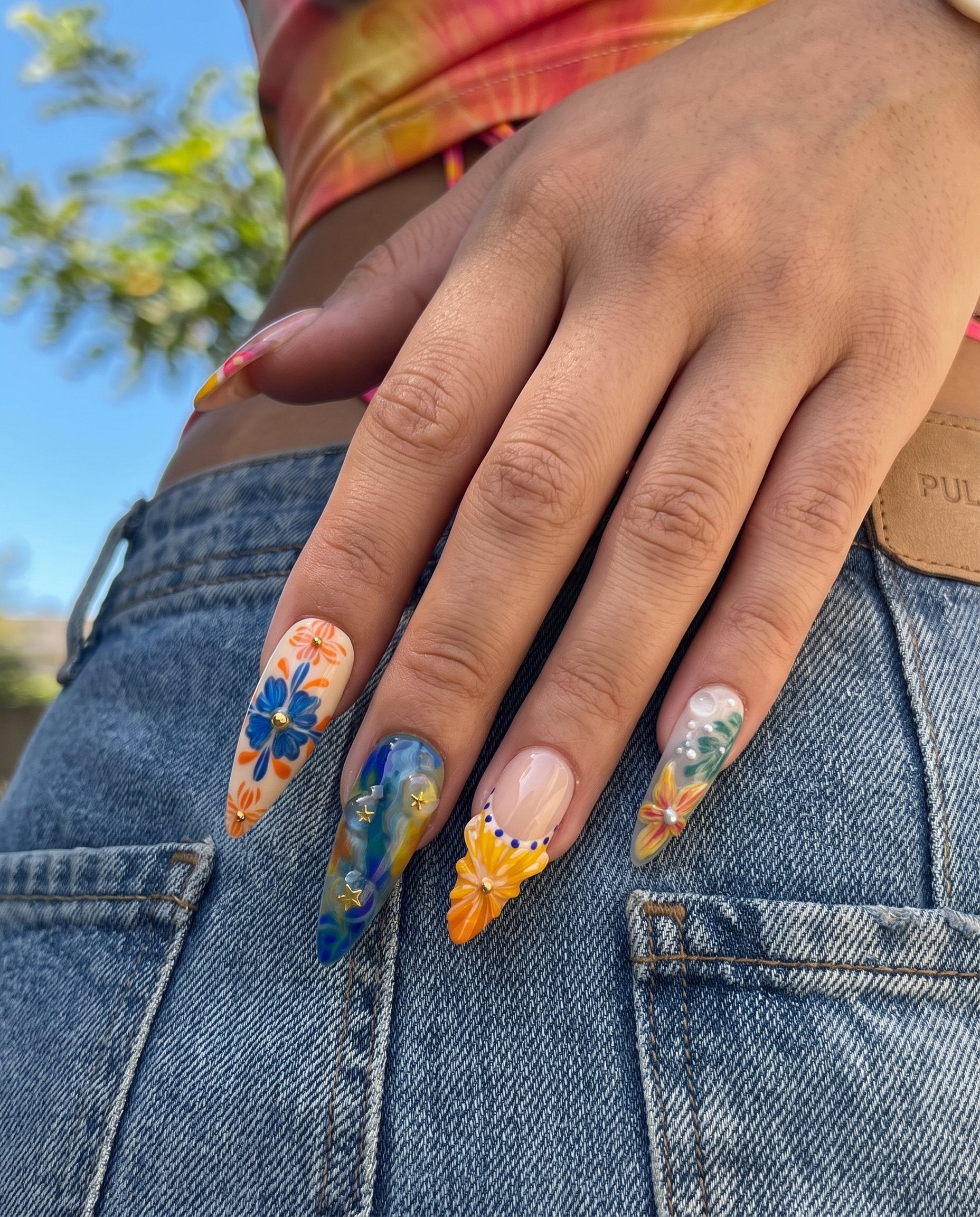 A set of ten stiletto-shaped press-on nails featuring a Mediterranean summer theme with 3D starfish, sun charms, yellow flowers, blue tile patterns, and ocean wave designs.