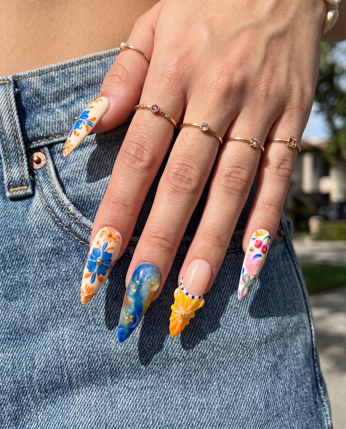 A set of ten stiletto-shaped press-on nails featuring a Mediterranean summer theme with 3D starfish, sun charms, yellow flowers, blue tile patterns, and ocean wave designs.