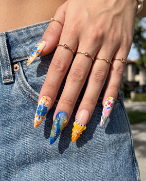 A set of ten stiletto-shaped press-on nails featuring a Mediterranean summer theme with 3D starfish, sun charms, yellow flowers, blue tile patterns, and ocean wave designs.