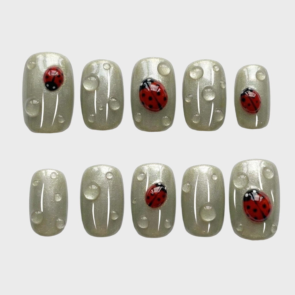 Sage green press-on nails featuring red ladybug motifs and clear dewdrop accents.